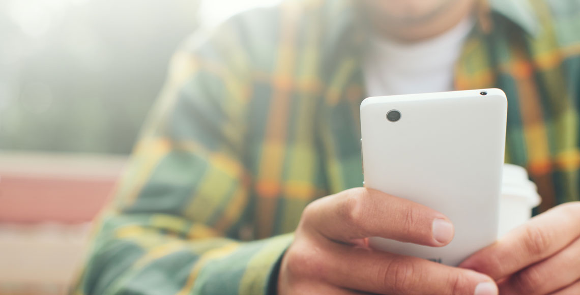 Young man with cup of coffee and smartphone in hand sitting in street cafe, browsing web
