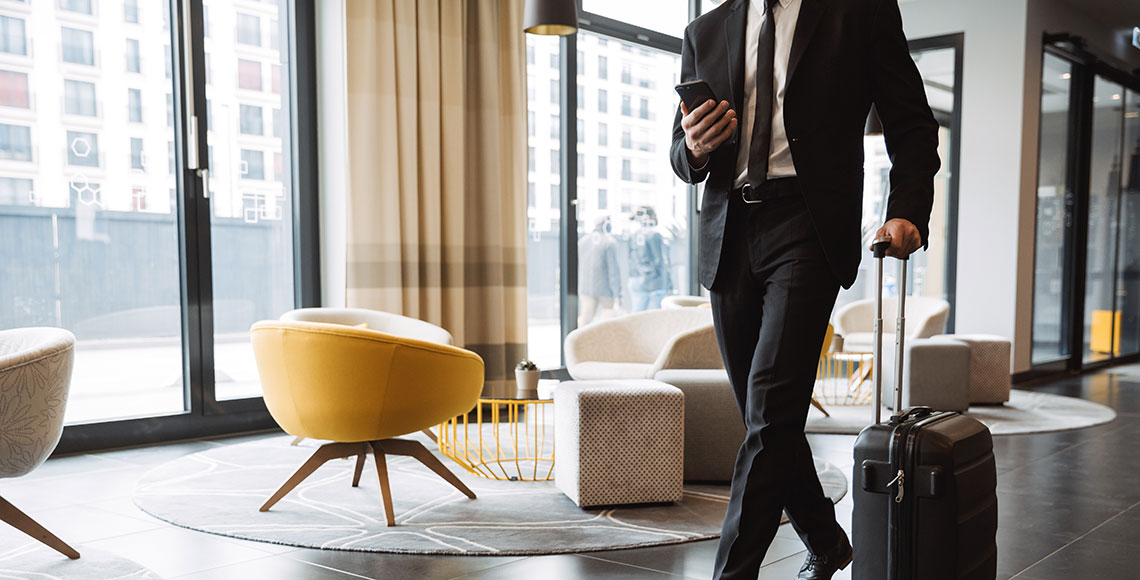 Cropped photo of successful businessman wearing suit holding smartphone and walking with suitcase in hotel lobby